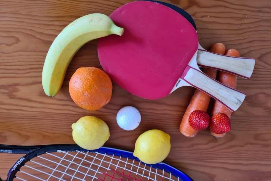 Gesamtthema und Ausdruck des Bildes: Gesundheit, Sport und gute Ernährung. Auf diesem Foto sind Oragnen, Zitronen, Karotten, Bananen und Erdbeeren zu sehen. Zudem sind ein Squashschläger, zwei Tischtennisschläger und ein Golfball abgebildet.